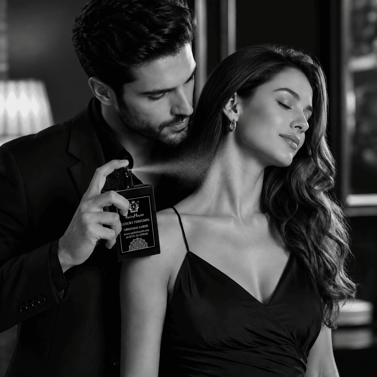 Black and white luxury perfume advertisement featuring a stylish man spraying RocheHayatt Obsessed Amber perfume on a woman in an elegant indoor setting, capturing romance, sophistication, and premium fragrance.